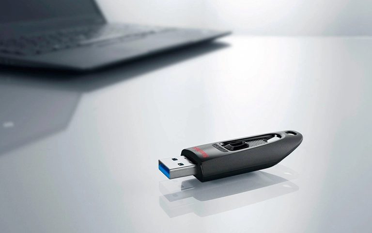Masa üzerinde duran siyah SanDisk Ultra USB bellek ve arkada dizüstü bilgisayar